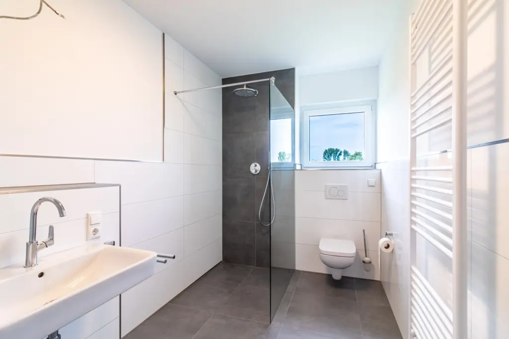 Moderner Badezimmer mit Dusche, Waschbecken und WC, helle Fliesen und große Fenster sorgen für viel Tageslicht.