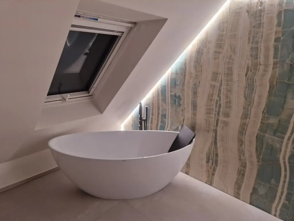 Freistehende Badewanne unter einem schrägen Fenster, umgeben von eleganter Wandverkleidung mit sanfter Beleuchtung.
