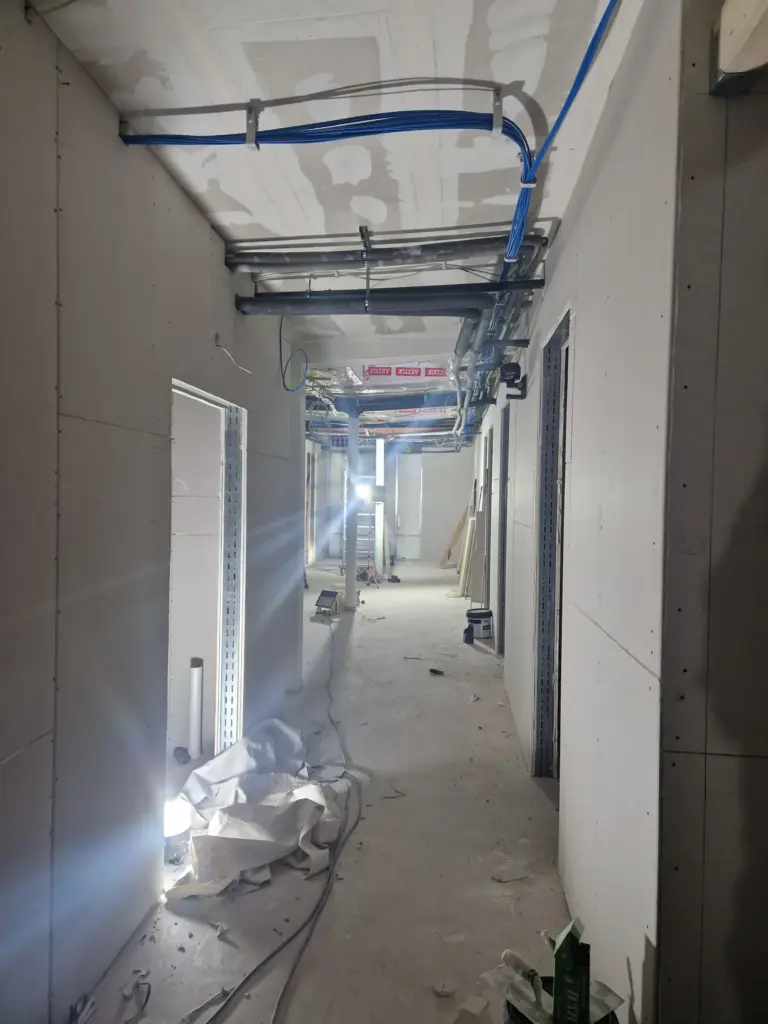 Bauarbeiten in einem unvollendeten Flur mit sichtbaren Rohren und Kabeln sowie Baustellenmaterialien auf dem Boden.