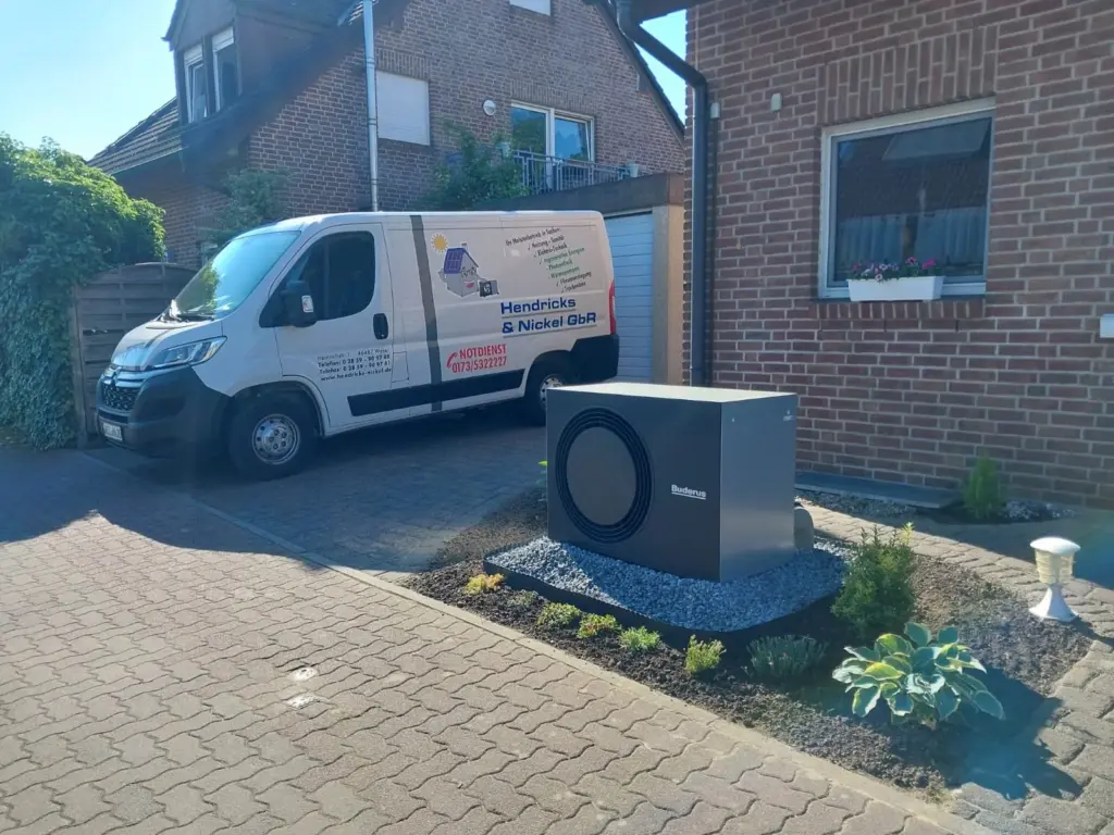 Ein Lieferwagen steht neben einer modernen Wärmepumpe in einer gepflasterten Auffahrt vor einem Wohnhaus.