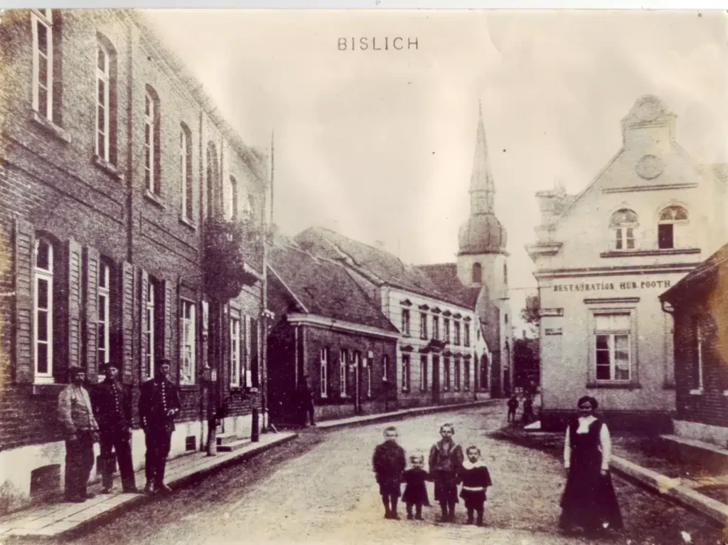 Historische Aufnahme einer Dorfstraße mit Häusern, Menschen und Kindern, einer Kirche im Hintergrund.