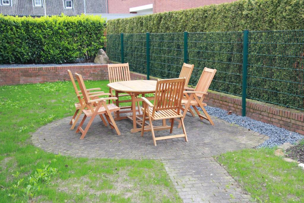 Ein runder Tisch mit fünf Holzstühlen steht auf einem gepflasterten Untergrund in einem Garten, umgeben von Grünhecken.