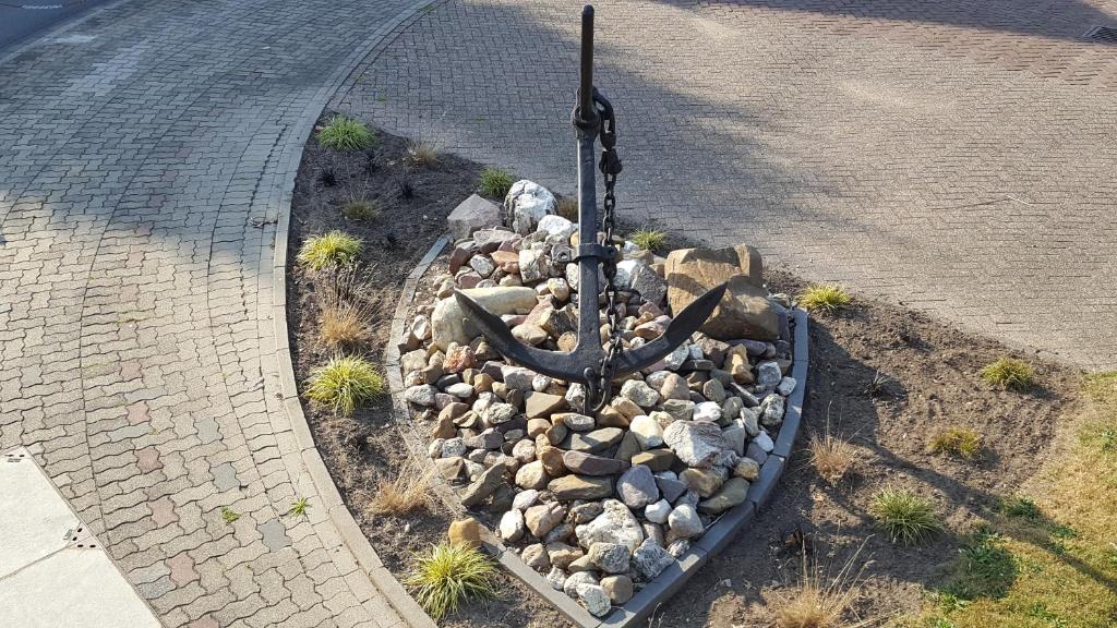 Ein großer Anker steht auf einem Kiesbett umgeben von Steinen und Gras in einer kleinen Pflanzfläche an einer Straßenecke.
