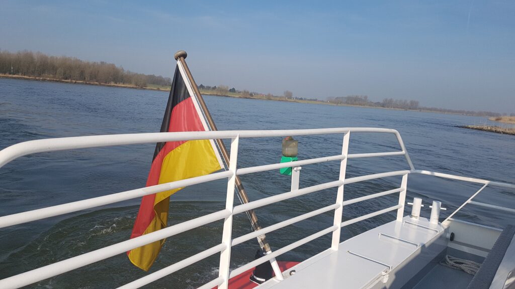 Ein Boot fährt auf einem Gewässer, im Hintergrund sind Bäume und eine deutsche Flagge weht am Heck.