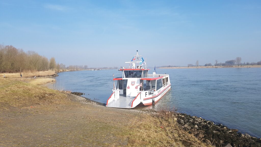 Ein Fahrgastschiff liegt an einem ruhigen Flussufer, umgeben von leicht bewaldeten Ufern und klarem Himmel.