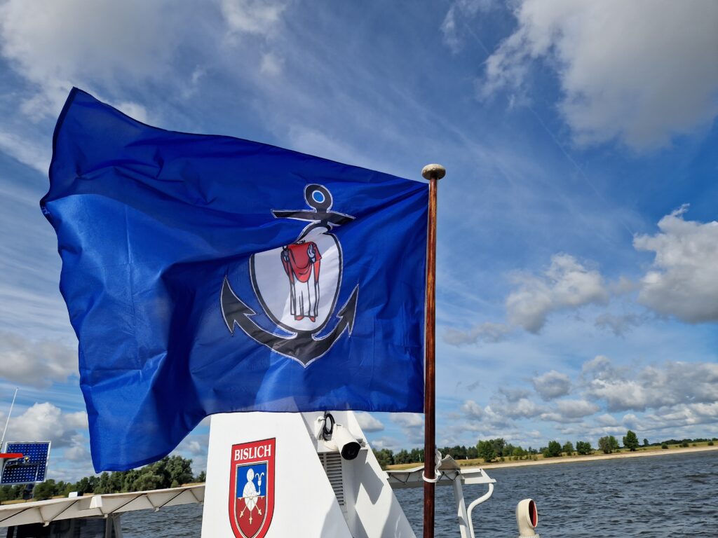 Eine blaue Flagge mit einem Anker und einer Gans weht vor einem wolkigen Himmel über einem Boot.