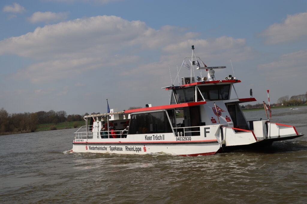 Ein Fahrgastschiff mit roter Markierung fährt auf einem Gewässer unter bewölkten Himmel.
