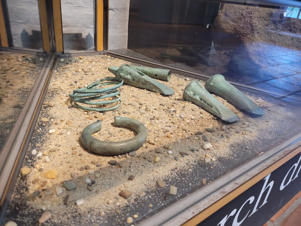 Vitrine mit antiken Bronzefunden, darunter Ringe und andere Artefakte, auf sandigem Untergrund ausgestaltet.