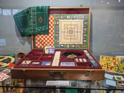 In einem Vitrinenschrank liegt ein kunstvoll gestaltetes Brettspiel mit bunten Spielfiguren und Spielkarten.