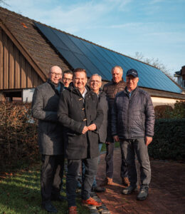 Sechs Personen stehen vor einem Haus mit Solaranlage auf dem Dach, umgeben von Garten und bei sonnigem Wetter.