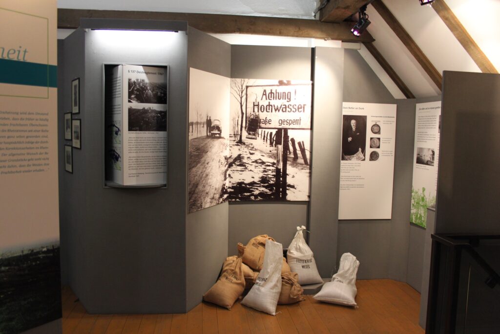 Eine Ausstellung zeigt historische Bilder und Infotafeln zum Thema Hochwasserschutz, umgeben von Sandsäcken.