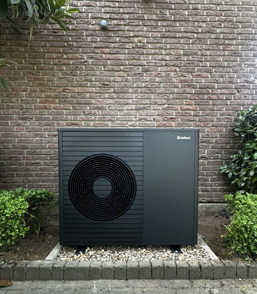 Energieeffiziente Wärmepumpe in einer Gartenlandschaft, umgeben von kleinen Pflanzen und einer Backsteinwand.