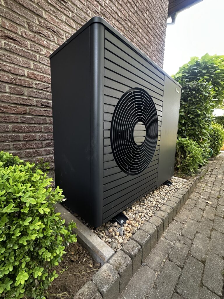 Eine schwarze Wärmepumpe steht neben einem gepflasterten Weg und einer Hecke an einer backsteinernen Wand.
