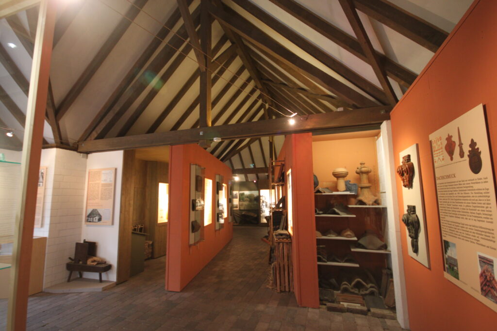Ein Innenraum eines Museums mit Holzbalkendecke, rot gestrichenen Wänden und Ausstellungsstücken in Vitrinen.
