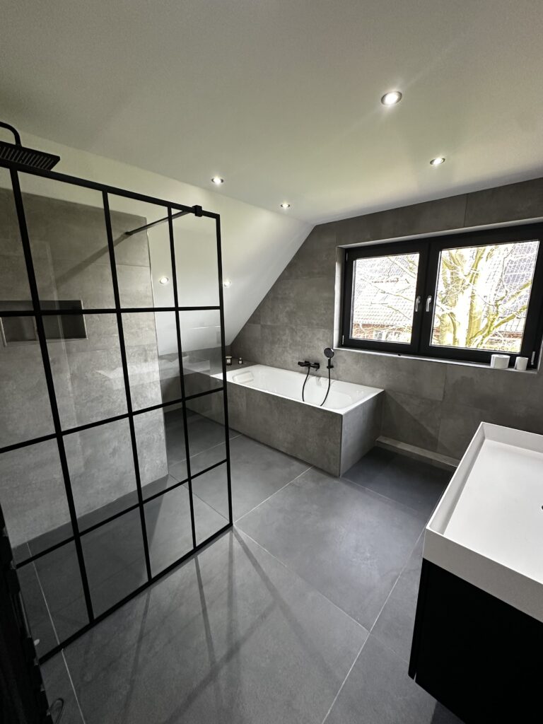 Modernes Badezimmer mit bodentiefer Dusche, freistehender Badewanne und großflächigen Fliesen in Grautönen.
