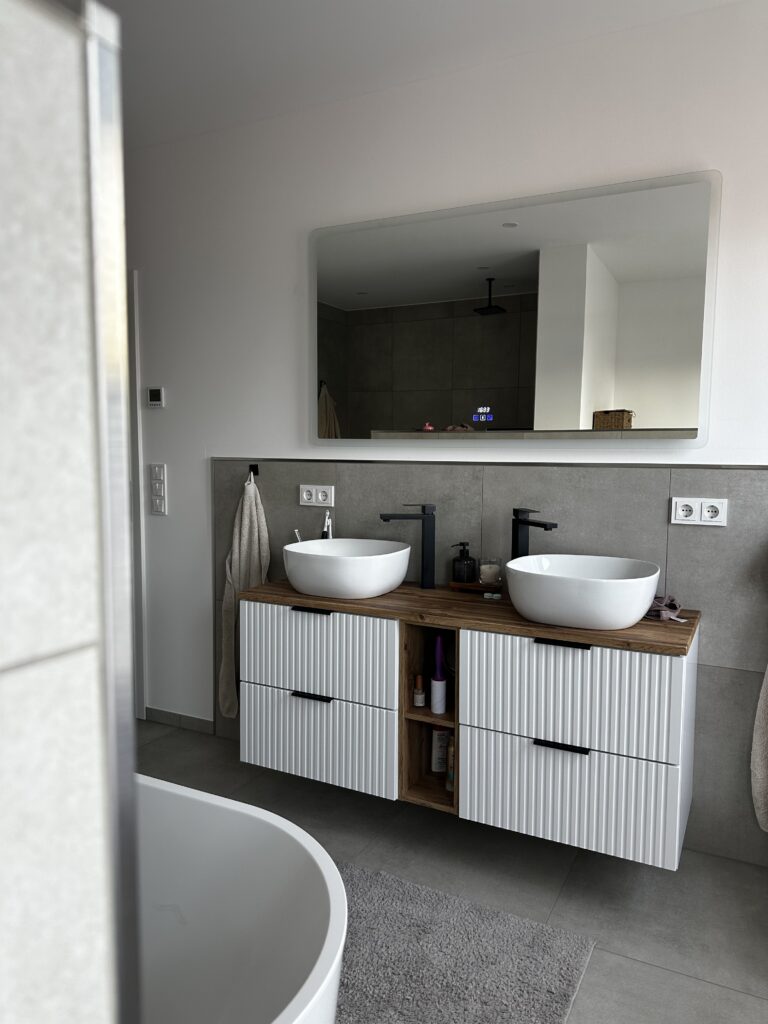Modernes Badezimmer mit zwei runden Waschbecken auf einem Holzregal, Spiegel darüber und Handtuchhalter an der Wand.