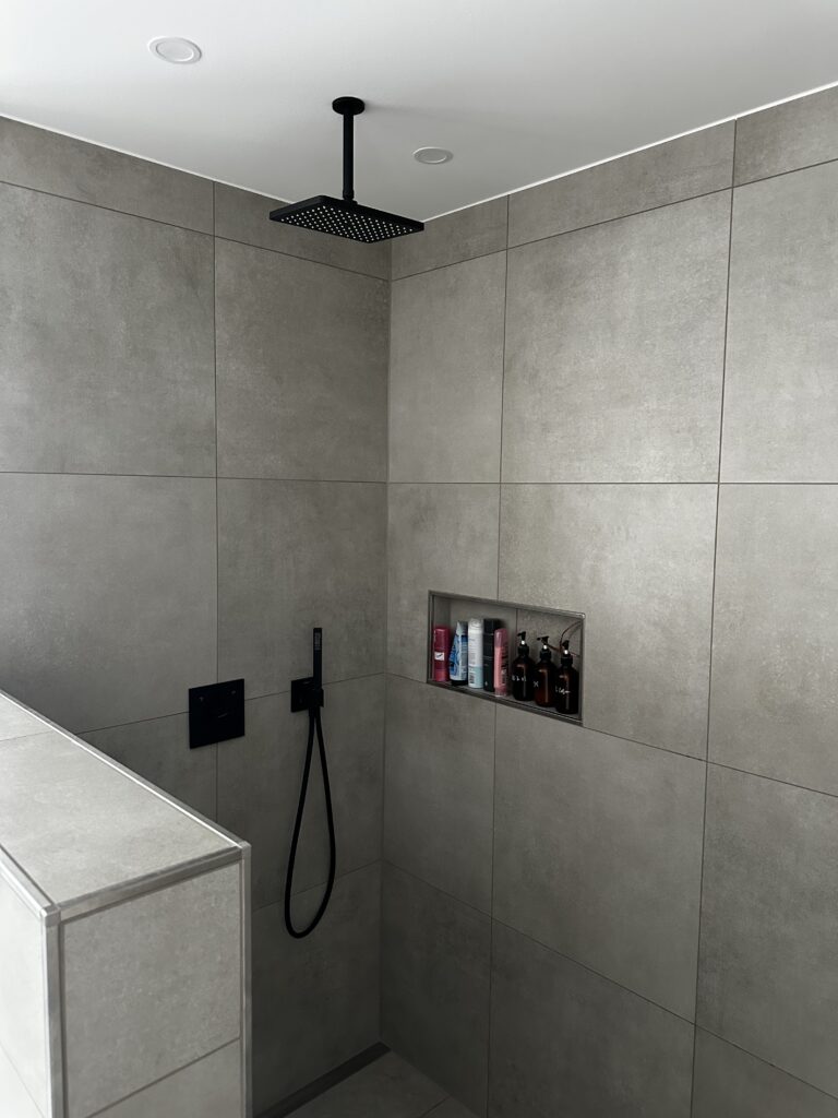 Moderne Dusche mit großem Regenbrausekopf und integrierter Nische für Pflegeprodukte auf grauen Fliesen.