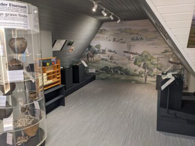 Ein Ausstellungsraum mit archäologischen Funden aus der Eisenzeit und einer Wandmalerei mit einer Landschaftsdarstellung.