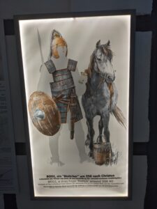 Darstellung eines kriegerischen Mannes um 550 nach Christus mit Rüstung, Schild und Pferd, daneben ein Eimer.