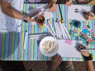 Mehrere Personen gestalten Puzzles und zeichnen mit Markern auf einem bunten Tisch, umgeben von Farben und Bastelmaterialien.