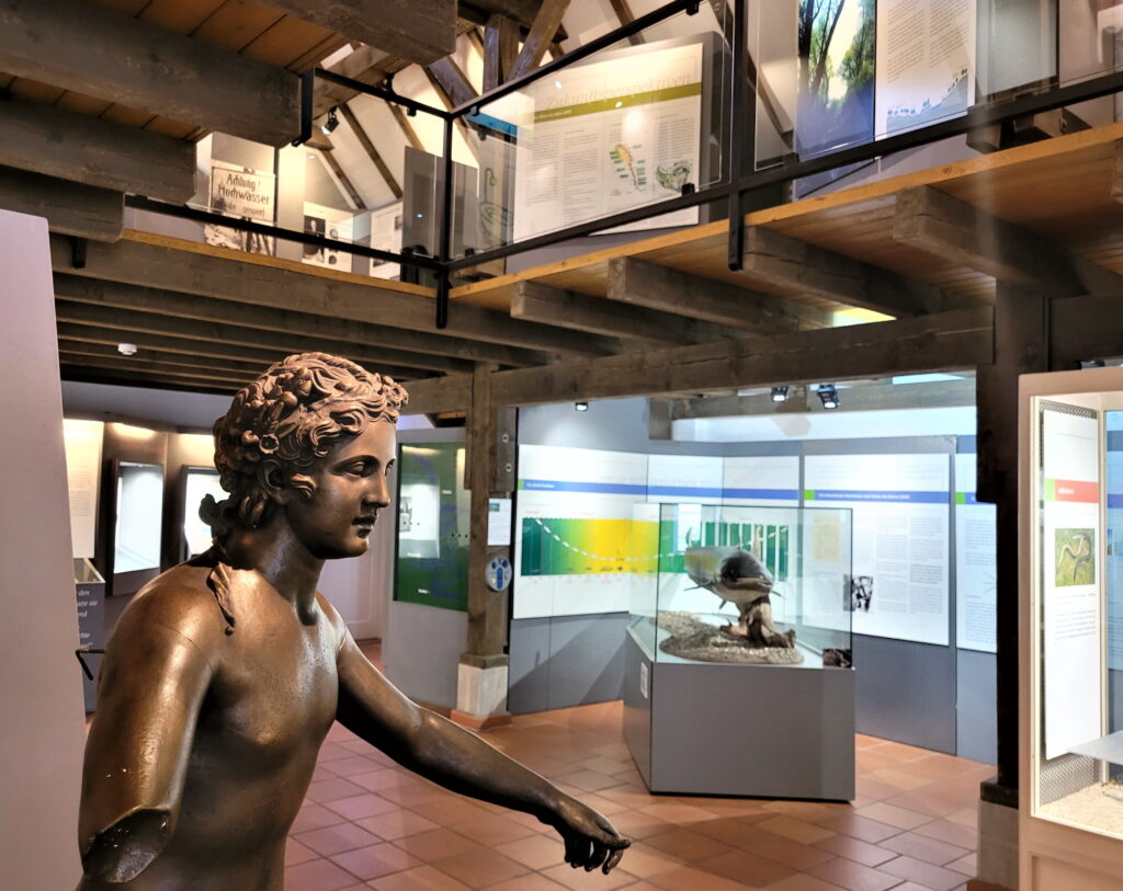 Eine bronzene Skulptur steht in einem modernen Ausstellungsraum mit Informationen und Vitrinen zur Natur und Kultur.