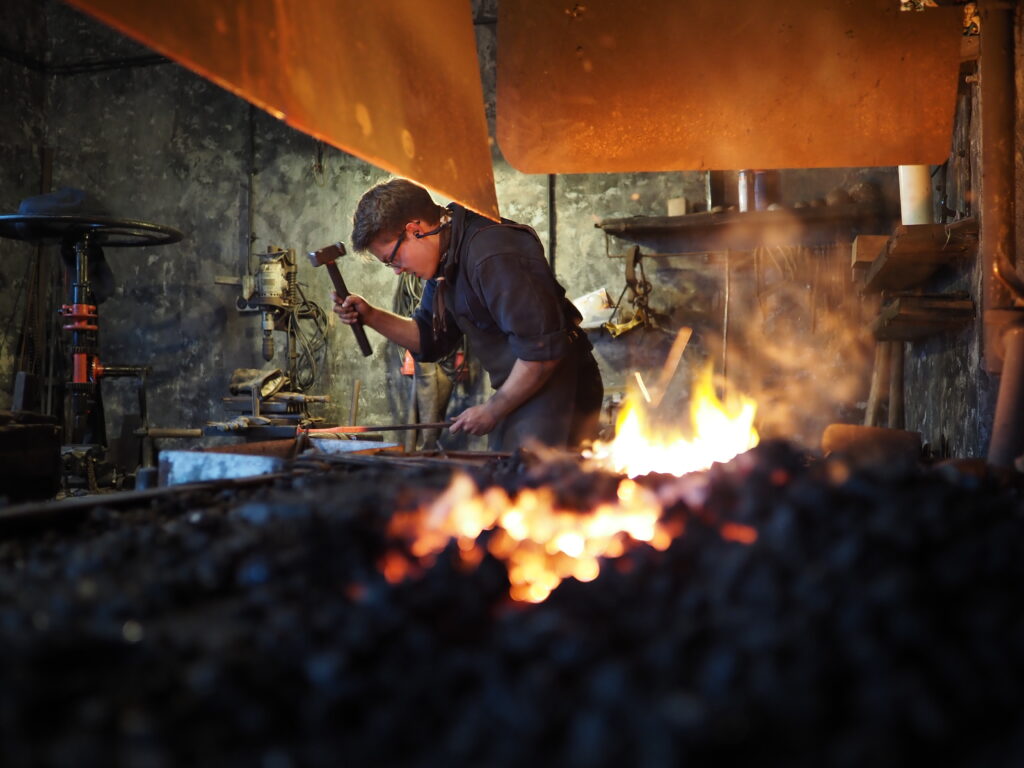 Ein Schmied arbeitet konzentriert am Amboss, während flammende Glut und Rauch aus dem Feuer hervorkommen.