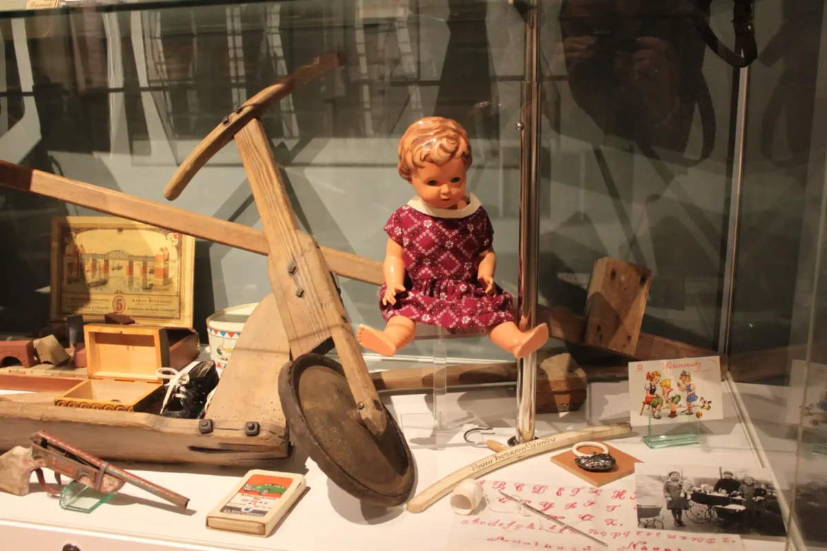 Eine alte Puppe sitzt auf einem Spielzeuglaufrad, umgeben von verschiedenen historischen Gegenständen und Fotografien.