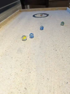 Auf einer sandigen Spielfläche rollen bunte Murmeln in Richtung eines Ziels in der Mitte.