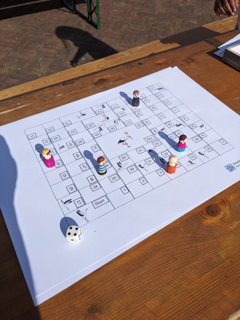 Auf einem Holztisch liegt ein buntes Spielbrett mit Figuren und einem Würfel, umgeben von ein paar Händen.