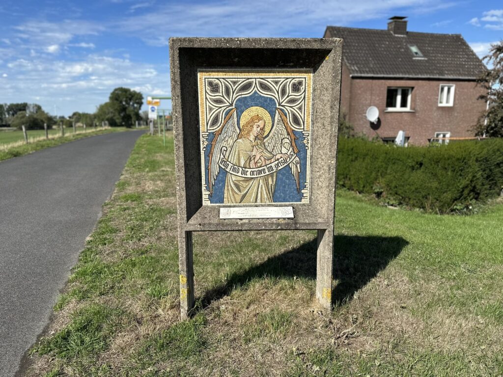 Ein dekoratives Steinrelief mit einer religiösen Darstellung steht am Straßenrand in einem ländlichen Gebiet.