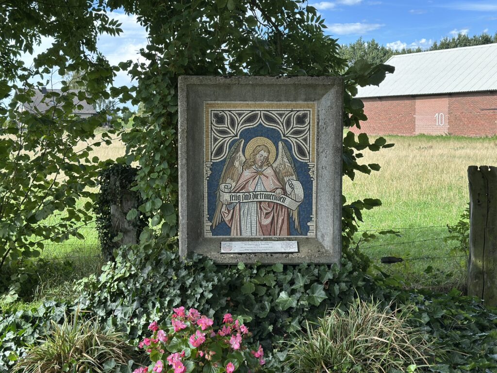 Ein mosaikartiger Engelsdarstellung ist von Pflanzen umgeben, im Hintergrund erstreckt sich eine Wiese und ein Hofgebäude.