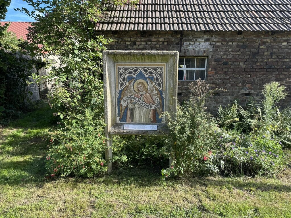 Ein Mosaik mit einem Heiligen steht vor einem alten Backsteingebäude, umgeben von Pflanzen und Blumen.