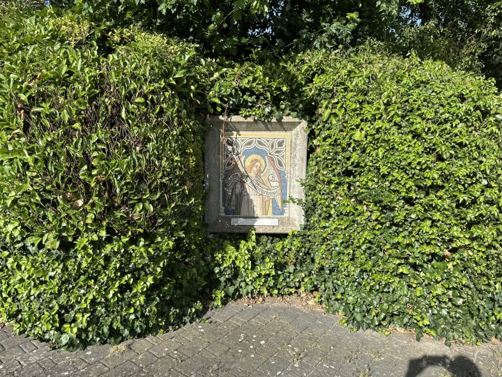 Ein mit Pflanzen umrandeter Bereich zeigt ein stilisiertes Wandbild, das in einer Hecke eingebettet ist.