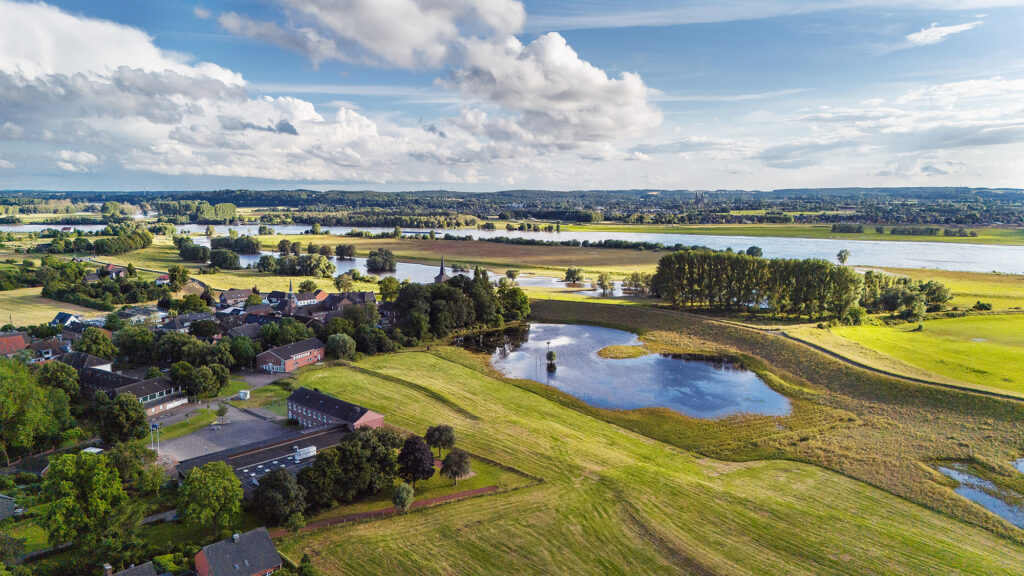 Landschaft mit Wiesen, einem Teich und verstreuten Wohnhäusern, umgeben von Bäumen und einem fließenden Gewässer.