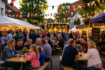 Ein belebtes Dorffest mit festlich dekorierten Ständen, Lichtern und einer Bühne, während Besucher miteinander feiern.
