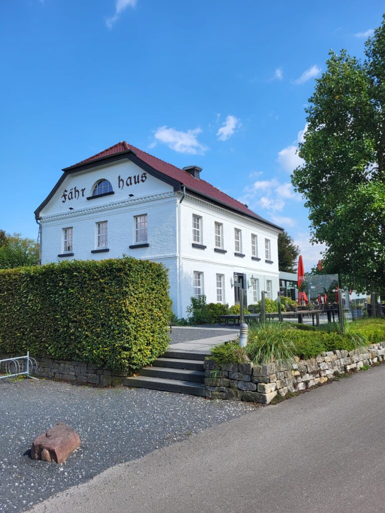 Weißes Gebäude mit rotem Dach und Schriftzug „Fährhaus“, umgeben von grünen Hecken und einem gepflasterten Weg.