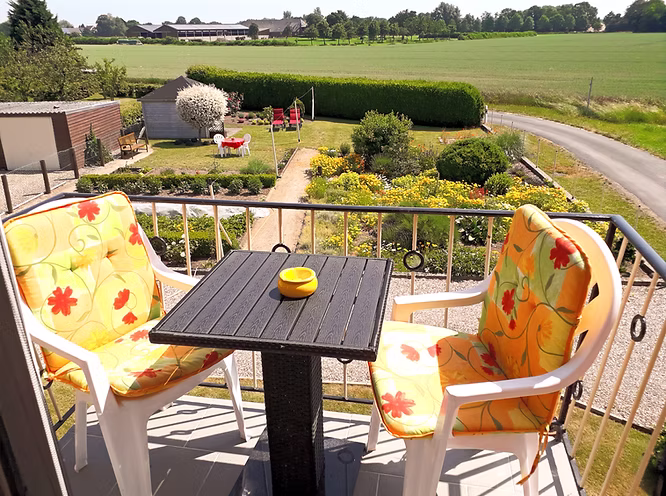 Ein kleiner quadratischer Tisch mit zwei Gartenstühlen auf einem Balkon. Die Aussicht geht über den Garten auf die offenen Felder und Wiesen Bislichs.
