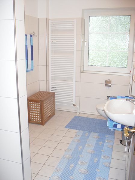 Helles Badezimmer mit Dusche, Waschbecken, Handtuchhaltern und einem Holzkorbschrank, dekoriert mit blauen Teppichen.