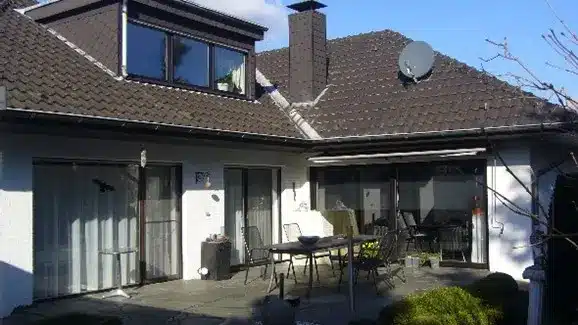 Helles Einfamilienhaus mit zwei Erkern, Terrasse und Gartenmöbeln unter blauem Himmel.