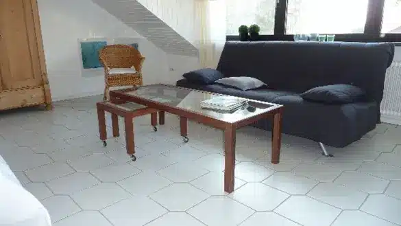Ein Wohnzimmer mit einem grauen Sofa, einem Glastisch und einem Korbsessel auf hellen Fliesen.