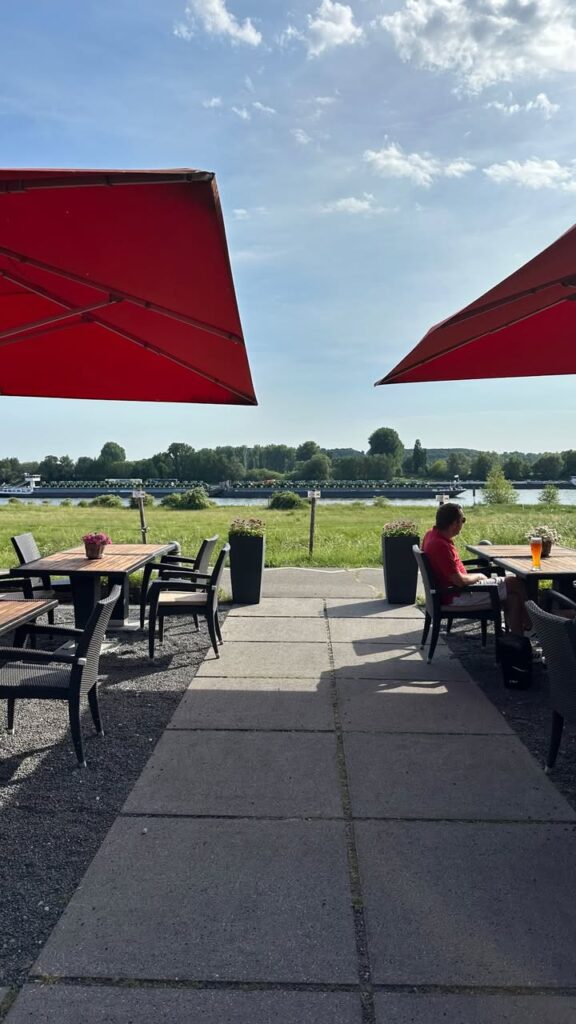 Blick auf eine Terrasse mit Gästen, roten Sonnenschirmen und Flusslandschaft im Hintergrund.