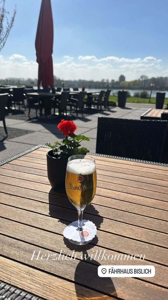 Ein Glas Bier steht auf einem Tisch mit blühenden Geranien im Freien, bei sonnigem Wetter am Fährhaus Bislich.