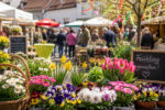 Bunte Blumenstände und Menschen auf einem lebhaften Frühlingsmarkt in einem malerischen Dorf.