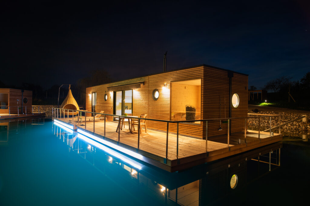 Ein modernes Holzhaus mit beleuchtetem Balkon steht nachts auf einem schwimmenden Fundament im ruhigen Wasser.