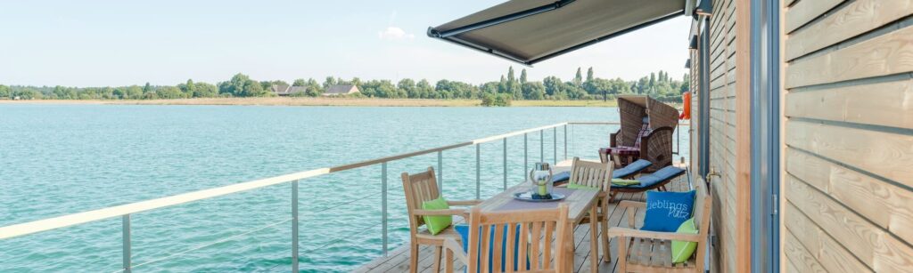 Holzterrasse mit Tisch und Stühlen am Wasser, im Hintergrund grüne Landschaft und ein paar Gebäude.