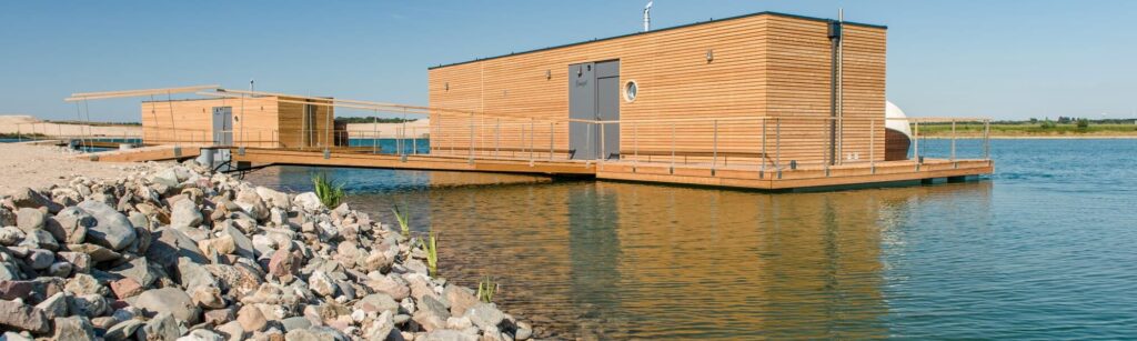 Moderne Holzhäuser auf einer schwimmenden Plattform im ruhigen Wasser, umgeben von Steinen und einem klaren Himmel.