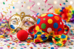 Bunte Clownshut, rote Nase, eine Maske und Konfetti liegen zwischen farbenfrohen Partydekorationen.