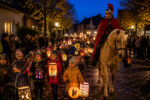 Eine festliche Laternenparade mit Kindern und einem Ritter auf einem Pferd in einer beleuchteten Dorfstraße bei Dämmerung.
