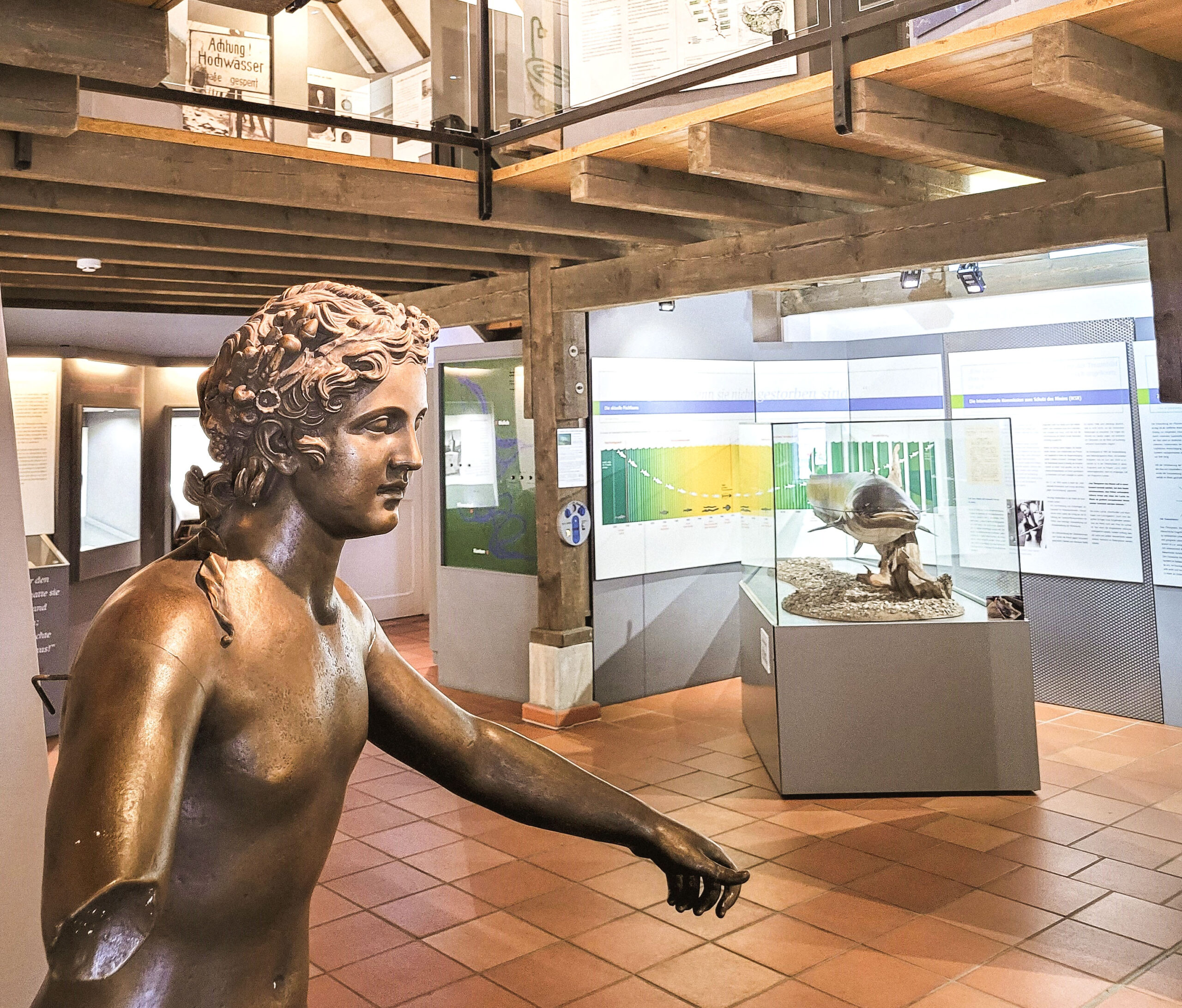 Eine bronzene Statue steht im Vordergrund eines Ausstellungsraums mit Informationswänden und Vitrinen im Hintergrund.