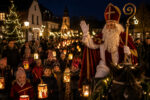 Eine festliche Menschenmenge mit Laternen begrüßt den Nikolaus auf einem Pferd in einer festlich beleuchteten Dorfstraße.